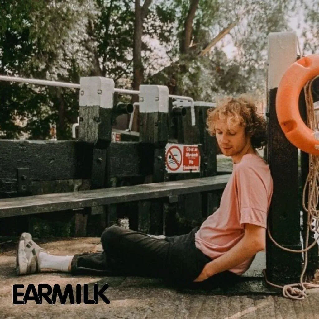 A young artist sitting by a canal lock, wearing a pastel pink shirt and black pants, with the EARMILK logo in the bottom left corner, representing music discovery and editorial features.