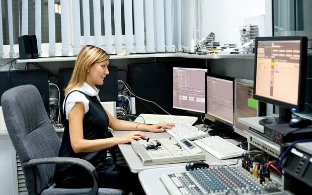 A female sound engineer working in a professional recording studio, surrounded by multiple monitors, an audio mixing console, and digital sound processing equipment.